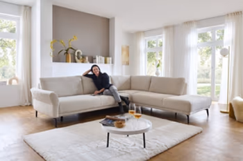 Auf dem Bild ist ein Wohnzimmer zu sehen. Eine helle, moderne Couch mit schwarzen Füßen. Es handelt sich hier um eine L Couch. Vor der Couch steht ein Couchtisch und darunter liegt ein schöner flauschiger, heller Teppich.